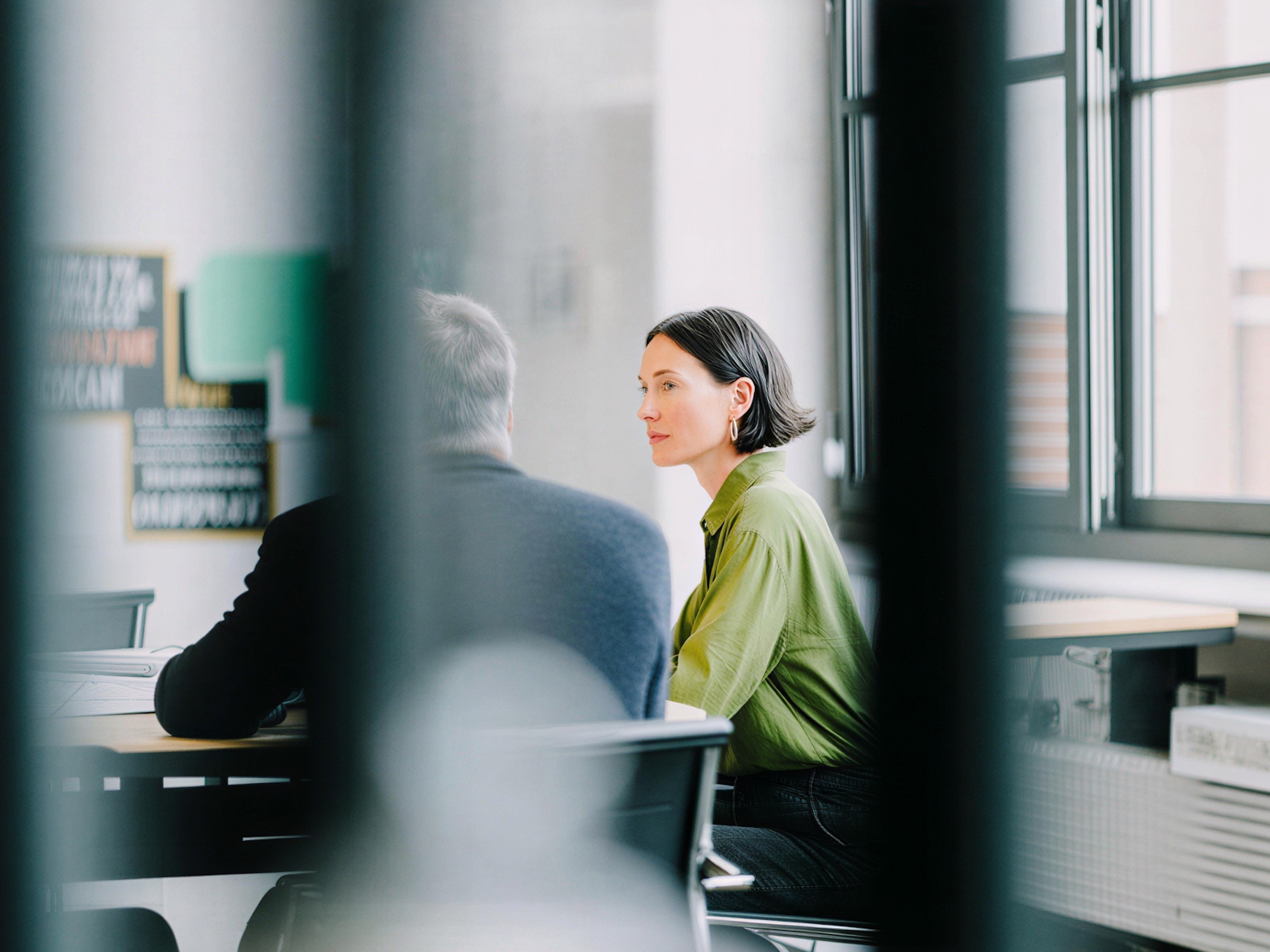 Two colleagues talking at a desk in an office, seen past a blurred column.